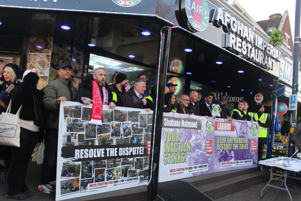 A landscape shot of striking workers and members of the community standing together outside the entrance to the Afghan Imperial Restaurant, holding a banner stating "resolve the dispute!", "your inaction stinks!" and "stop the rubbish, restart the talks". Some people look serious, some smile.