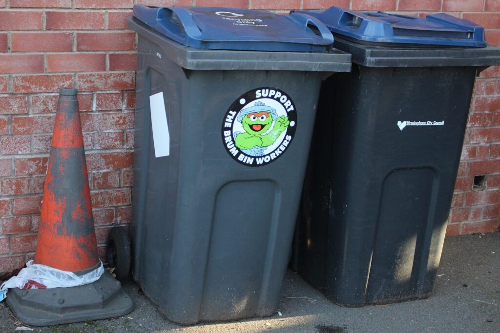 A traffic cone, to the left, is next to a recycling bin which has a "support the Brum bin workers" sticker on it's front, next to a general waste bin which has "Birmingham City Council" written on the front.