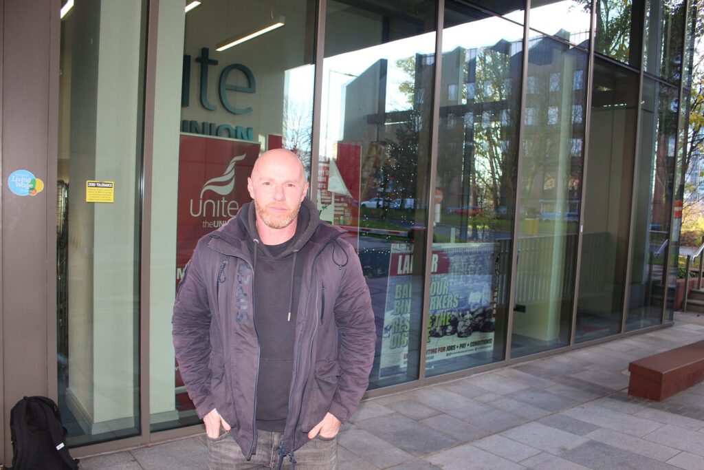 A portrait of Matt Reid outside of the Unite the Union reception window. He looks serious and stands with his hands tucked into his front pockets. There are banners in the window promoting bin worker industrial action.