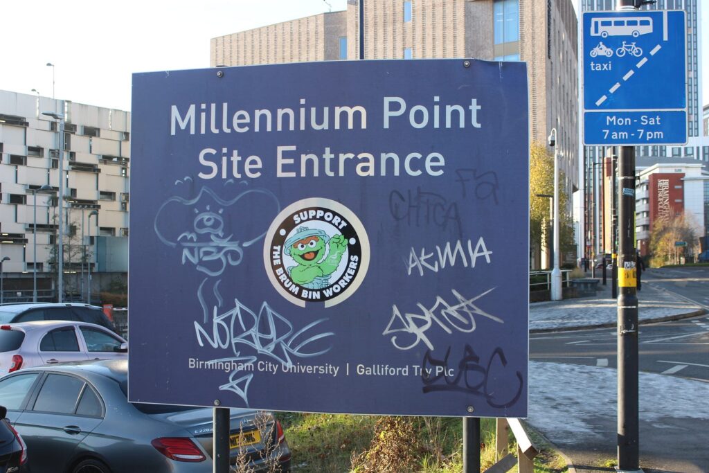 A "Millennium Point Site Entrance" sign, covered in graffiti but bearing a "support the Brum bin workers" sticker in the middle.