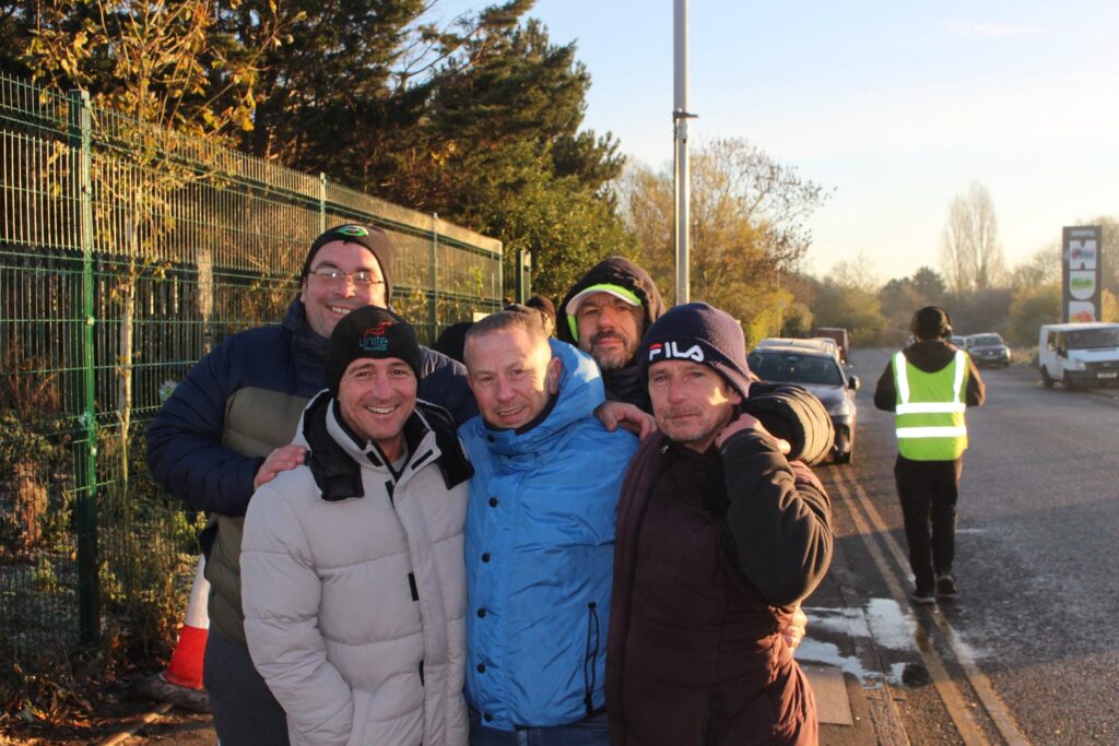 Striking workers huddle and smile in a shot together on the picket line. They are dressed warmly for the freezing weather - one worker wears a Unite the Union hat.