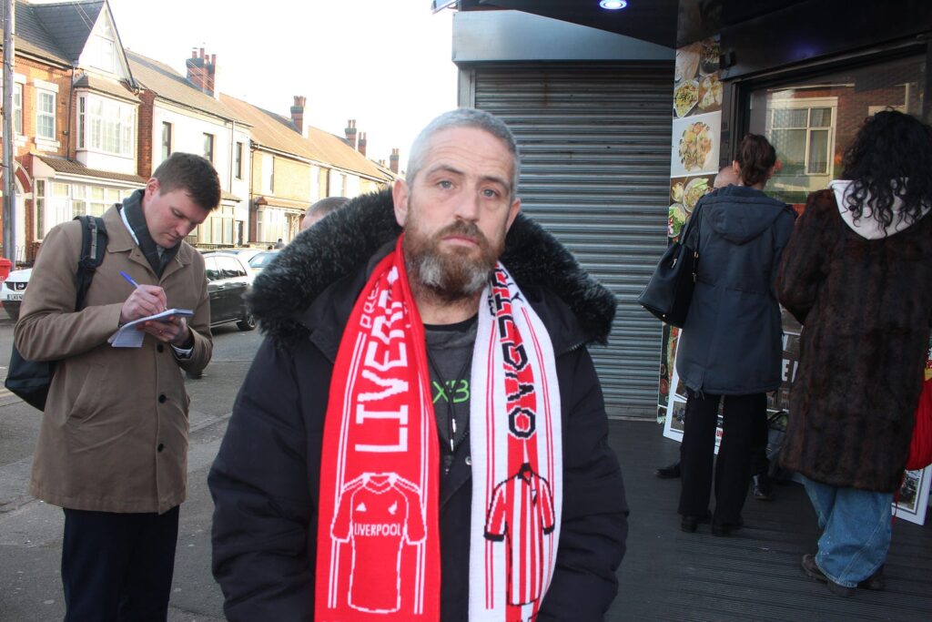 A portrait of Sam Yates on the street corner outside of the restaurant. He look serious. He wears a Liverpool football club scarf.