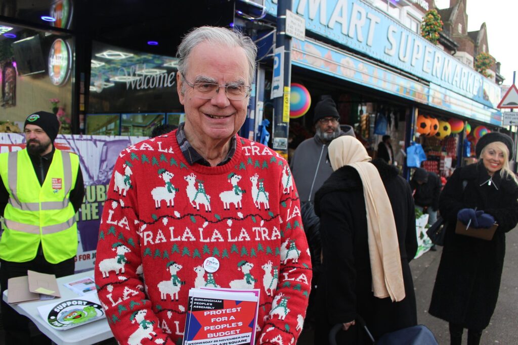A portrait of Stuart Richardson, smiling outside of the Afghan Imperial Restaurant, wearing his red and green Christmas jumper and holding a 'protest for a people's budget' flyer.