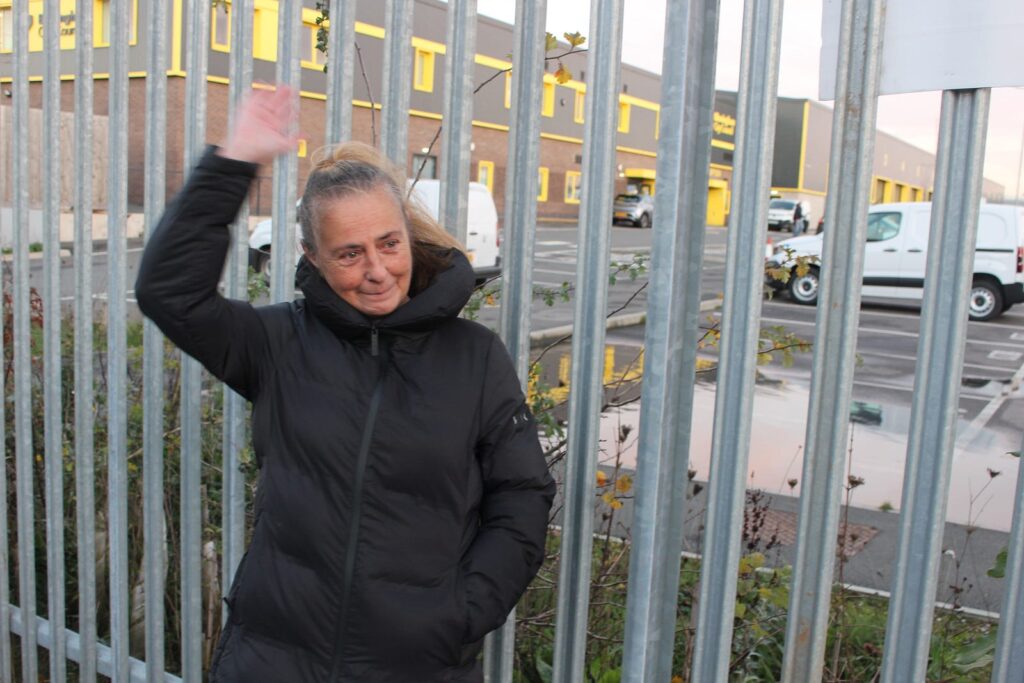 Wendy stands in front of the Tyseley depot gates, waving at a car driving past behind the camera.