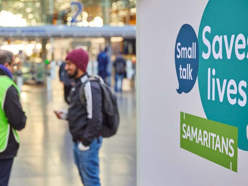 A Samaritans volunteer talking to a passenger in front of a Small Talks Saves Lives sign