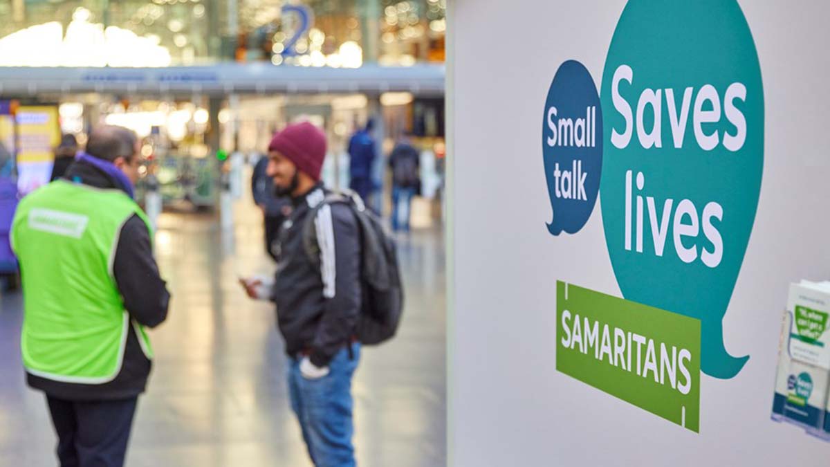 A Samaritans volunteer talking to a passenger in front of a Small Talks Saves Lives sign